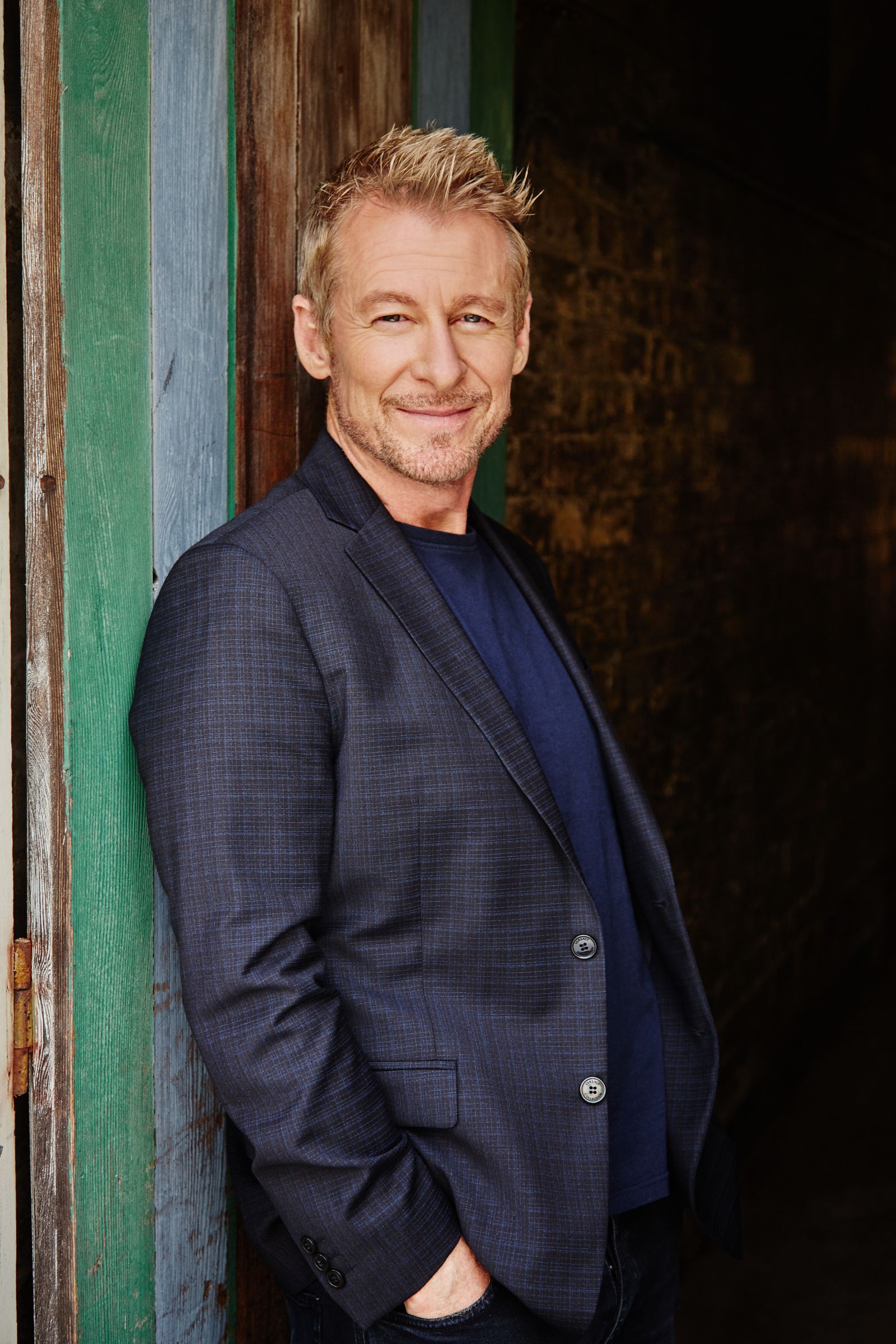 Smiling man in navy blazer leans against rustic doorway, hands in pockets, editorial portrait.